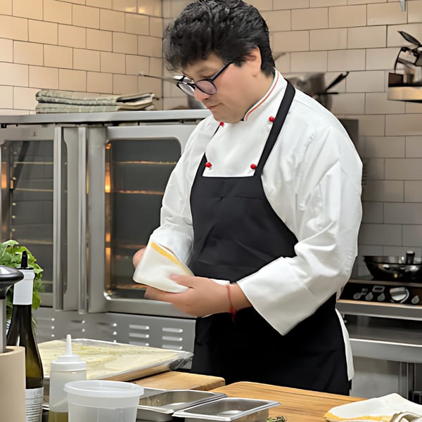 Chef in a white coat and black apron carefully folding a white cloth in a professional kitchen.