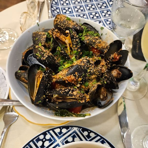 A white bowl of mussels topped with breadcrumbs and parsley, served on a restaurant table.